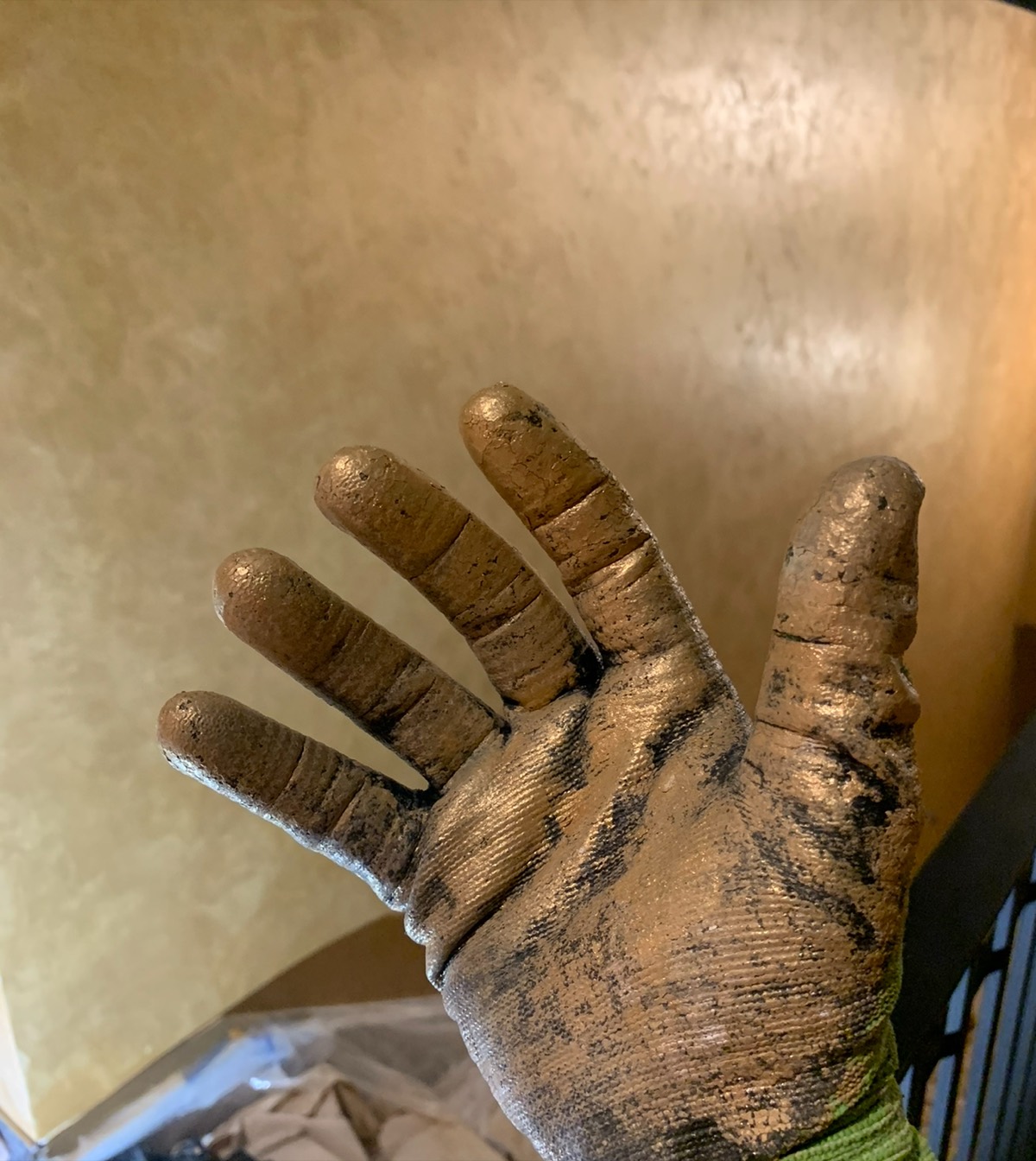 A gloved hand coated in gold plaster, held up in front of a freshly finished metallic plaster wall