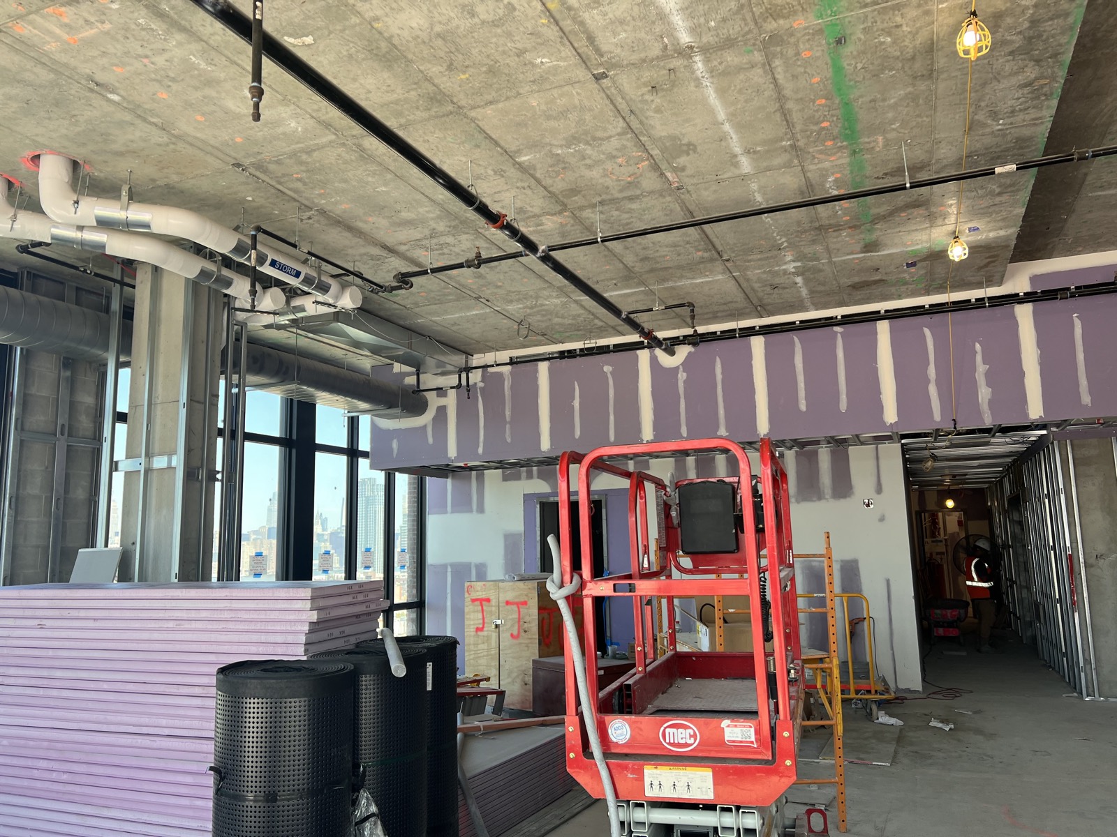 An interior job site in progress: taped drywall, a scissor lift and stacked boards, with the city skyline through the windows